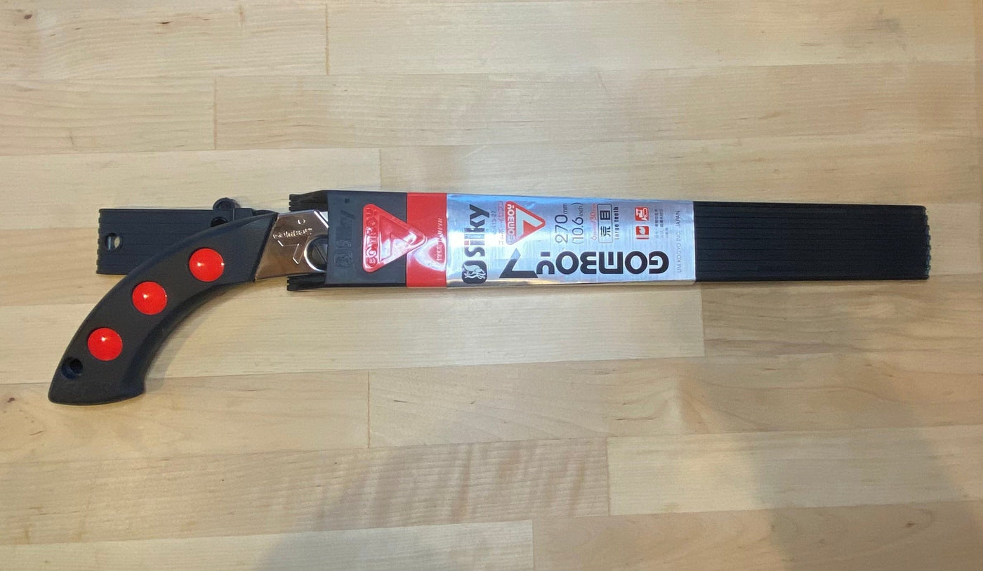 A black-handled SHT Silky Gomboy 270 folding saw with red grips, essential for timber framing, is displayed in its packaging on a light wooden surface. The packaging shows the SHT brand name and product details.
