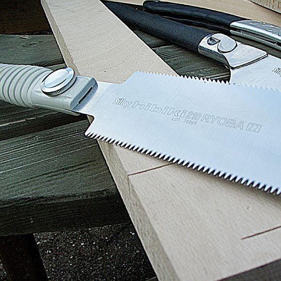 A close-up of the SHT Silky Hibiki Ryoba Handsaw with a white handle and impulse-hardened teeth rests on a wooden plank, while other hand tools are visible in the background on a workbench.