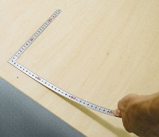 On a wooden surface, a hand holds one end of the SHWA Traditional Japanese Flexible Carpenter Square, a tool etched with metric and inch graduations, bent at a right angle.