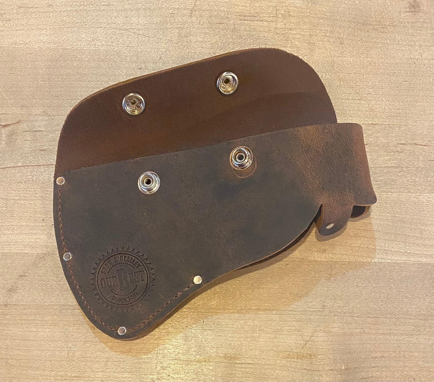 A brown leather pouch with metal fasteners, shown open and laid flat on a wooden surface. A circular embossed logo is visible on one side. Perfect for outdoor enthusiasts, it's designed to carry high-quality tools like the SNO Snow & Nealley Hudson Bay Camping Axe.