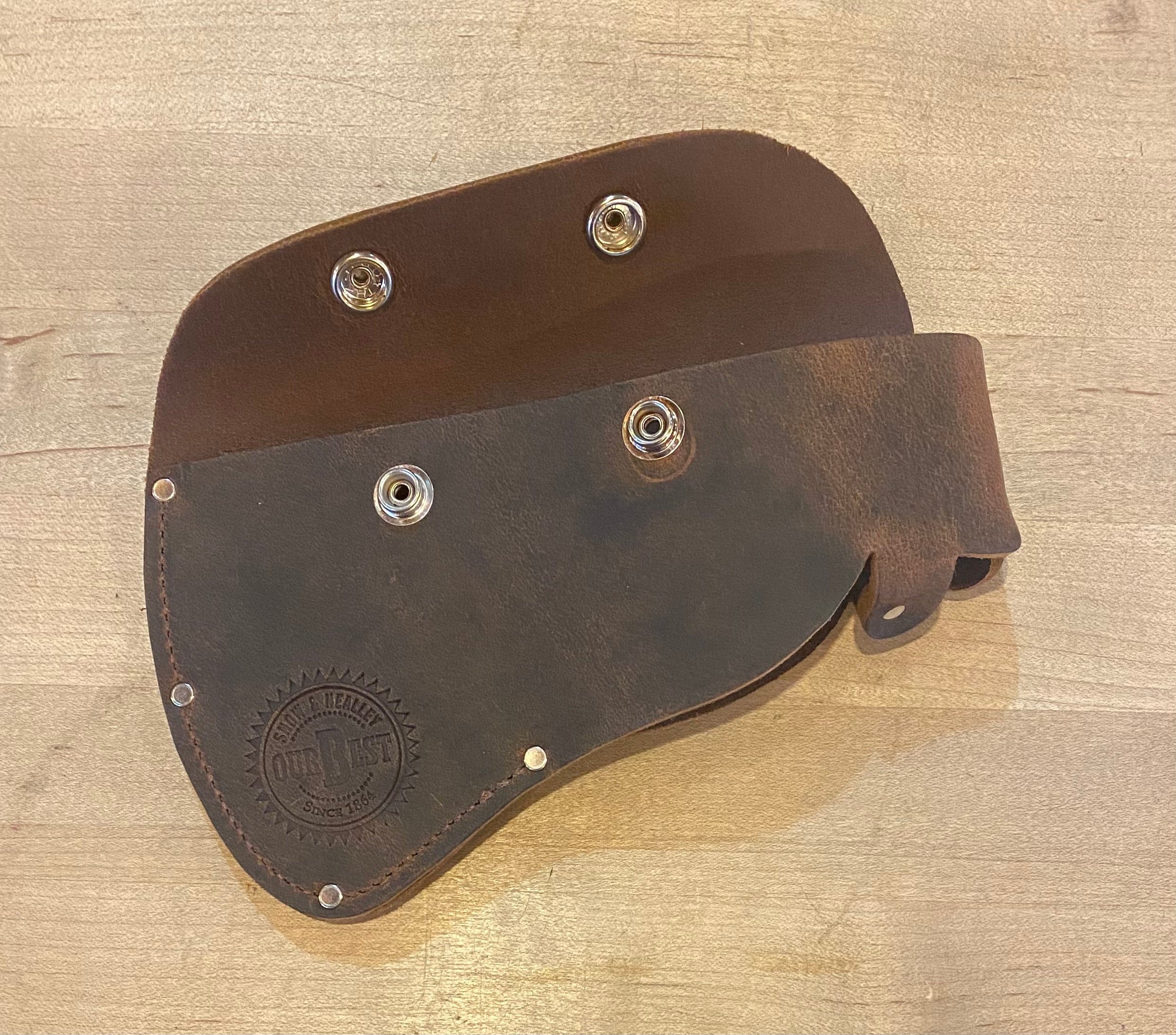 A brown leather pouch with metal fasteners, shown open and laid flat on a wooden surface. A circular embossed logo is visible on one side. Perfect for outdoor enthusiasts, it's designed to carry high-quality tools like the SNO Snow & Nealley Hudson Bay Camping Axe.