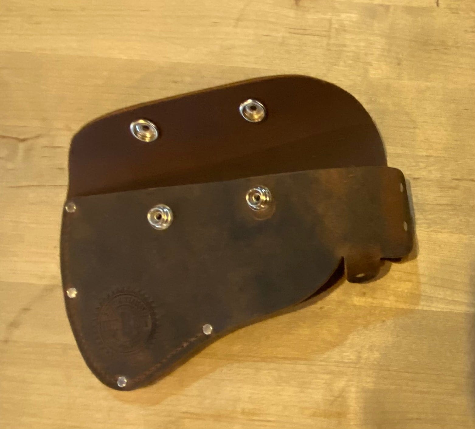 A brown leather SNO Campers Belt Axe holster with metal snap buttons rests on a light wooden surface. Slightly worn, it features an embossed circular logo—perfect for carrying your axe on hiking and camping trips.