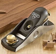 The 9-1/2 Block Plane Sweetheart Bailey Line by STN, a small hand plane for woodworking with an adjustable mouth, featuring a metal body and a brass adjustment knob, sits on a workbench surrounded by wood shavings. Its precision-ground cast iron base ensures smooth operation.