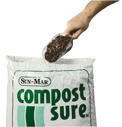 A hand holding a scoop of composting bulking material above a white bag labeled "SUN Compost Sure," suggesting fast decomposition for use in your Sun-Mar Composting Toilet.