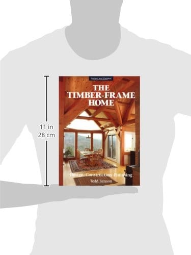 A person holds "The Timber-Frame Home 2nd Edition" by Taunton Press, showcasing the elegance of timber framing. The 11-inch tall book features a captivating image of a timber-framed interior on its cover.