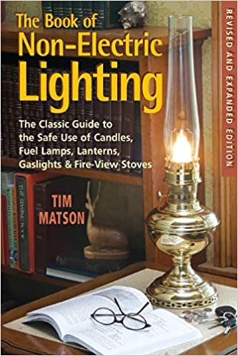 A vintage brass oil lamp glows on a wooden desk beside an open copy of The Book of Non-Electric Lighting: The Classic Guide to the Safe Use of Candles, Fuel Lamps, Lanterns, and Gaslights & Fire-View Stoves by W. W. Norton & Company.
