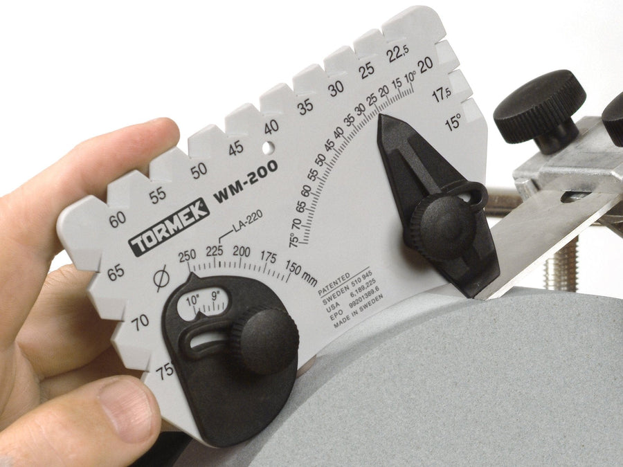 Close-up of a TOR Tormek Angle Master being adjusted by a hand. The tool measures angles on sharpening equipment, showing various degree markings, a knob for fine adjustments, and accommodates grindstone diameter to ensure precise edge angle settings.