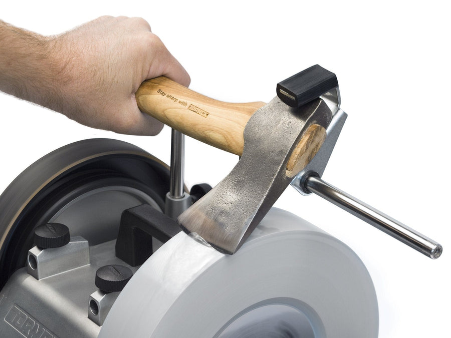 A hand holds an axe being sharpened on a rotating stone, guided precisely by the TOR Tormek Axe Jig to maintain consistent grinding pressure.