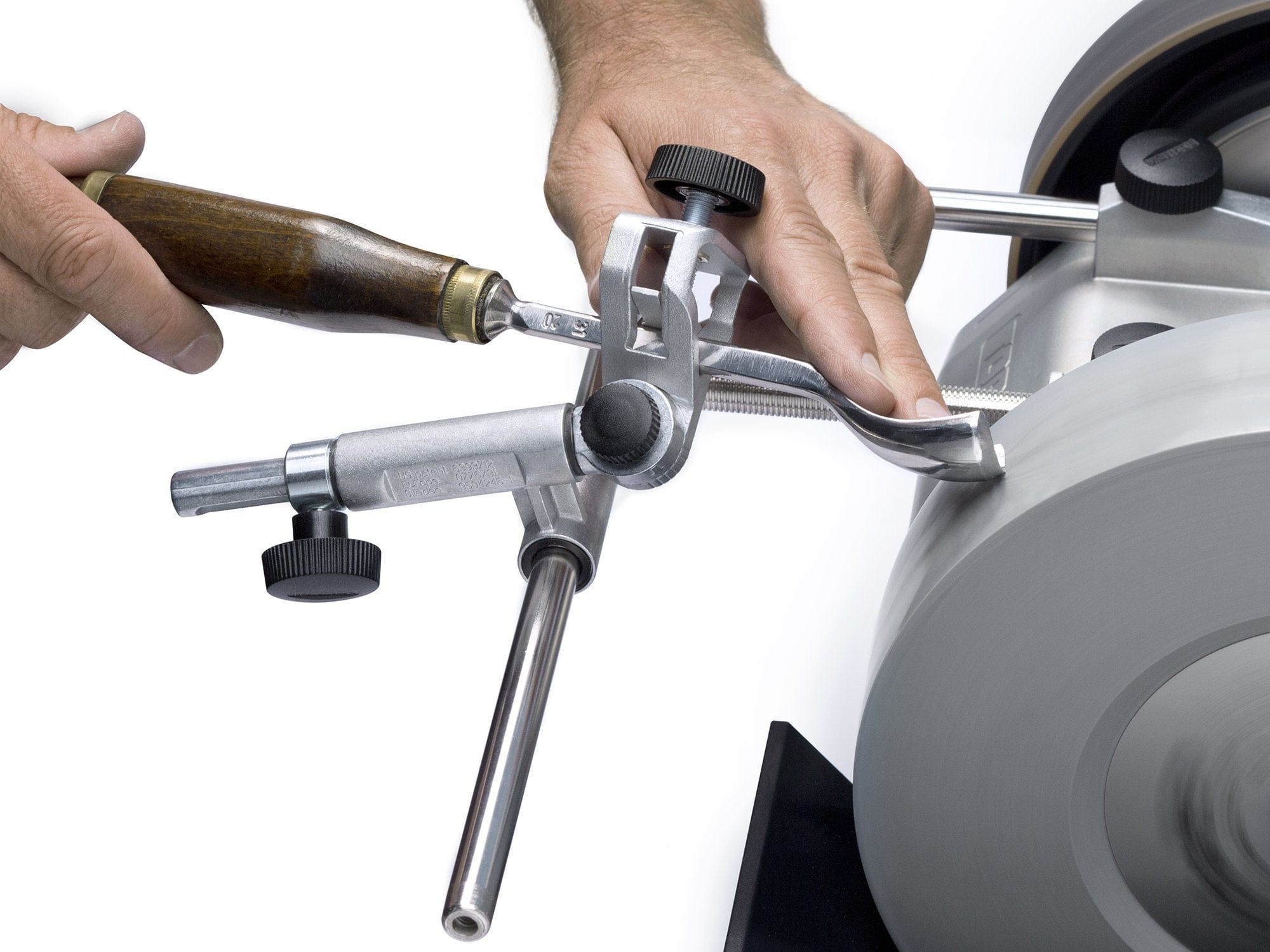 Close-up of hands sharpening woodturning tools on a rotating grinding wheel using the TOR Tormek Gouge Jig, showing adjustment knobs and visible metal parts.