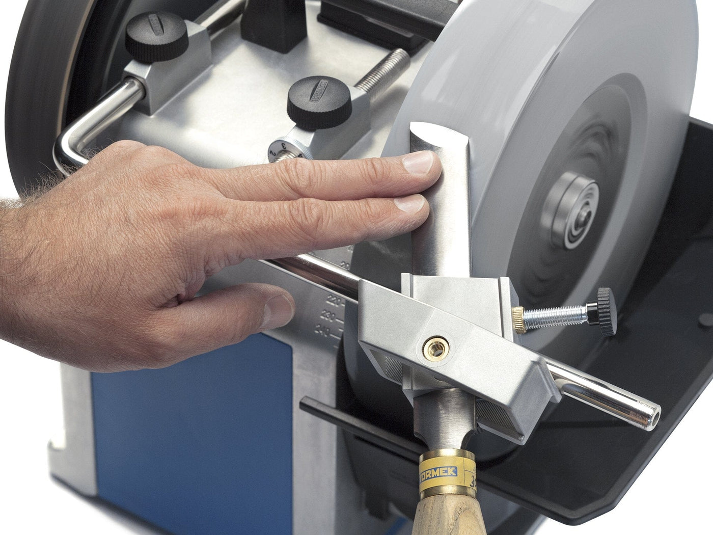 A person's hand touches a square chisel being sharpened using the TOR Tormek Multi Jig on an electric grinding machine.