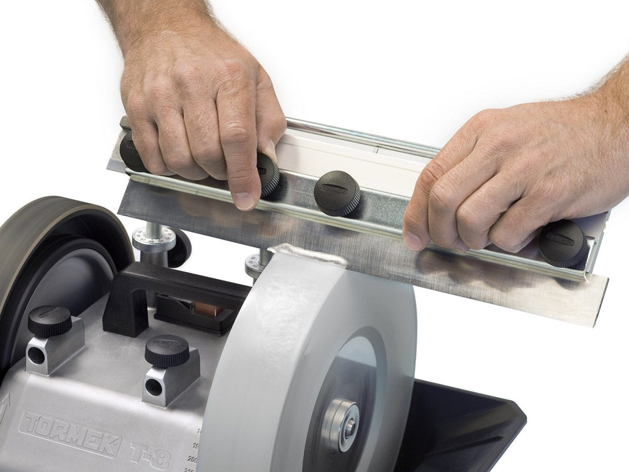Hands holding a blade against a rotating sharpening wheel on a TOR Tormek T-8 sharpening machine, using the Tormek Planer Blade Attachment for precision.
