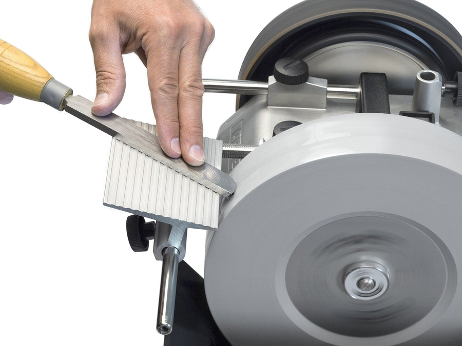 A person's hand is sharpening a chisel on a grinding wheel with a wooden handle, using the TOR Tormek Tool Rest to maintain the correct angle.