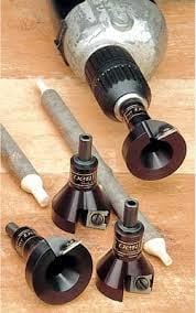 The Power Tenon Cutter by Veritas, featuring an aluminum body and three bits, is presented on a wooden surface with a power drill and two pencils. Ideal for crafting rustic furniture, this VER set ensures precision in every detail.