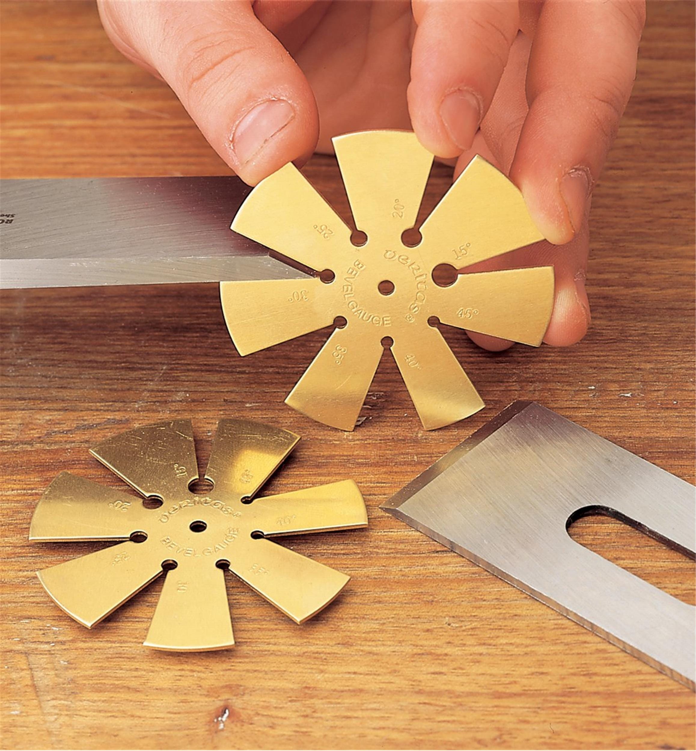 A hand holds a VER Veritas Bevel Gauge with engraved numbers beside a similar bevel gauge and two steel blades on a wooden surface.