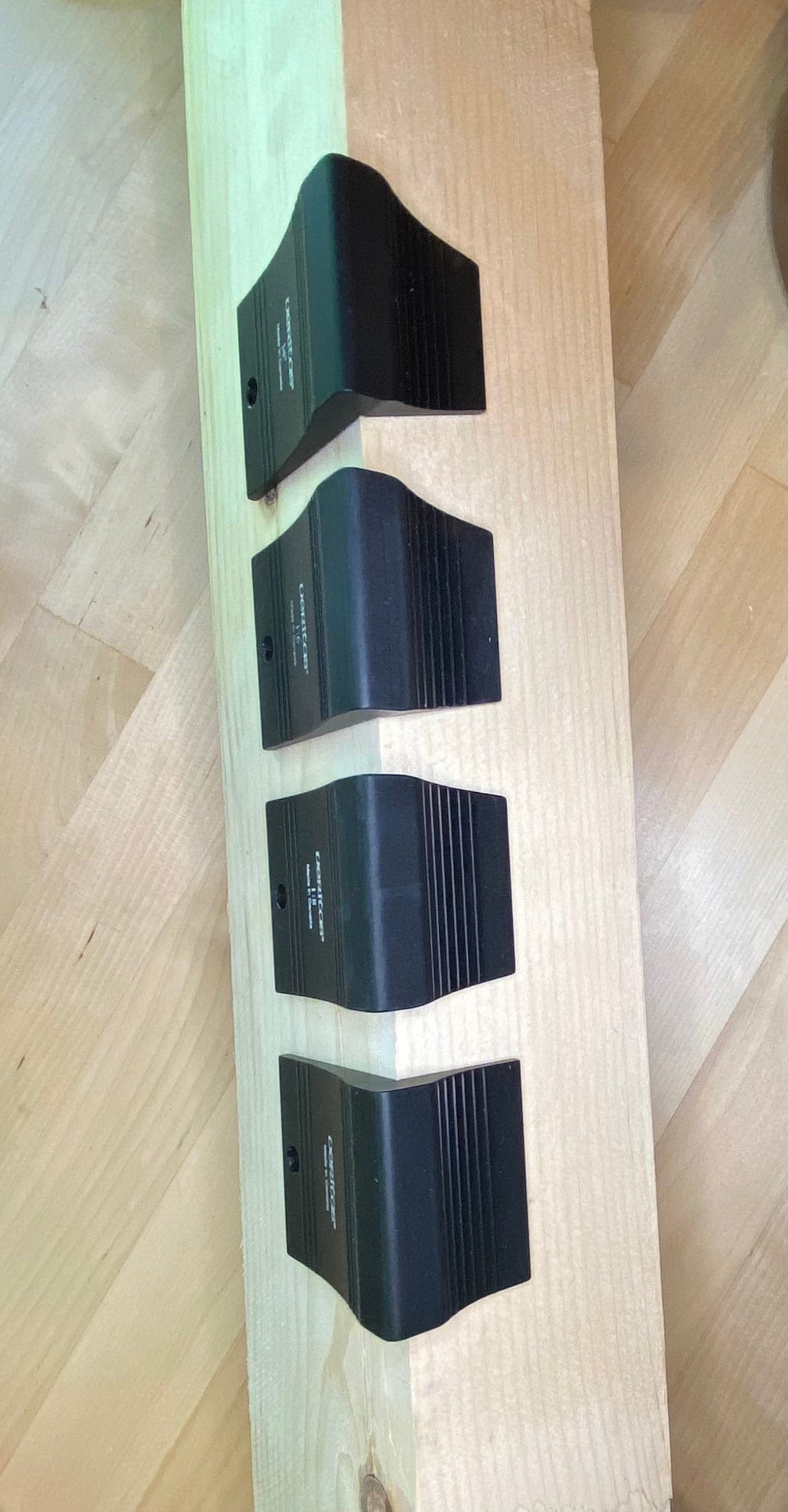 Four precision machined brackets from the VERITAS DOVETAIL SADDLE MARKER collection by VER, crafted in black metal, are mounted on a vertical wooden beam and placed on a light wood floor.