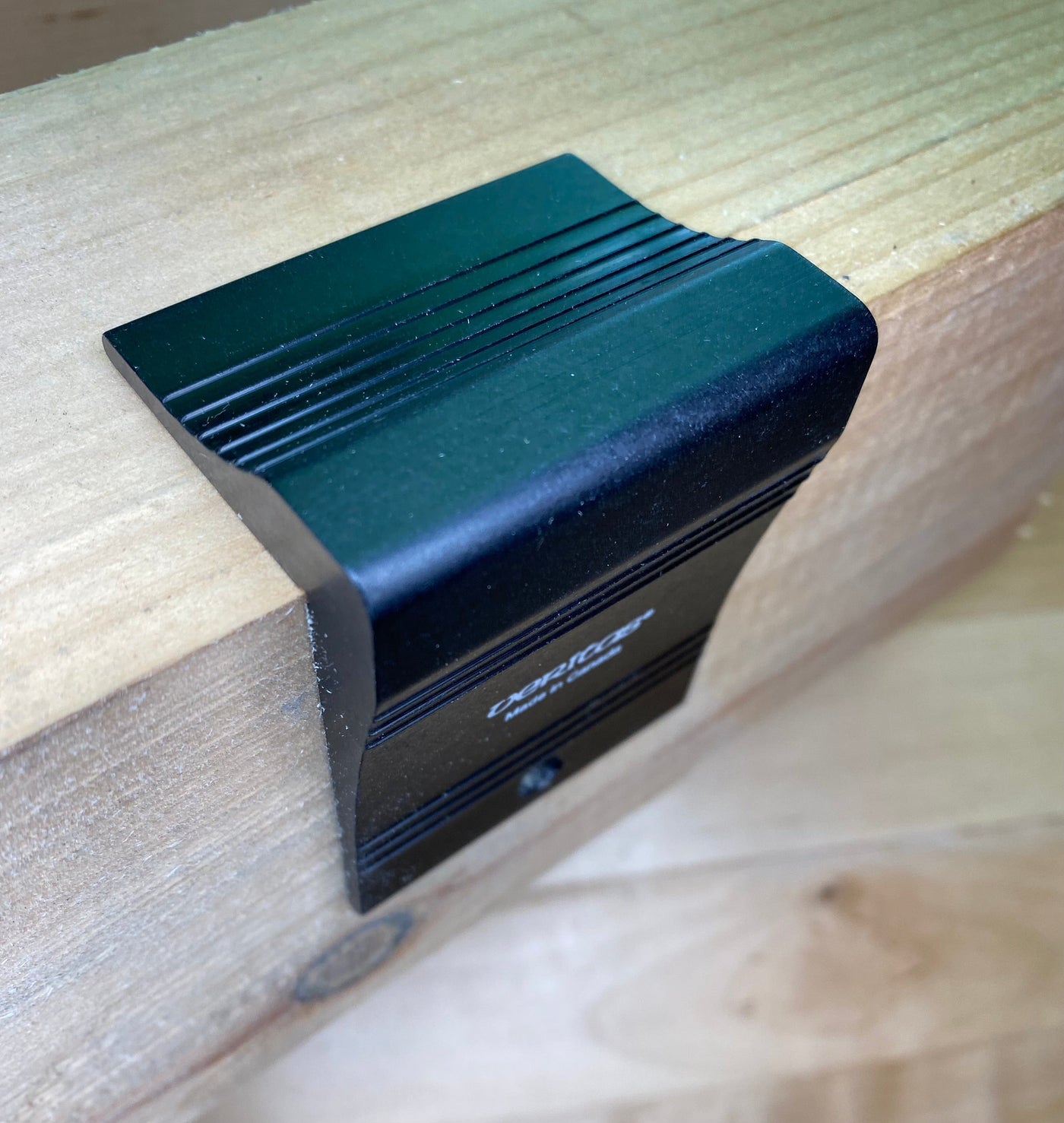 The VERITAS DOVETAIL SADDLE MARKER by VER features an anodized aluminum clip that securely attaches to the edge of a wooden surface, offering a sleek and durable hold.