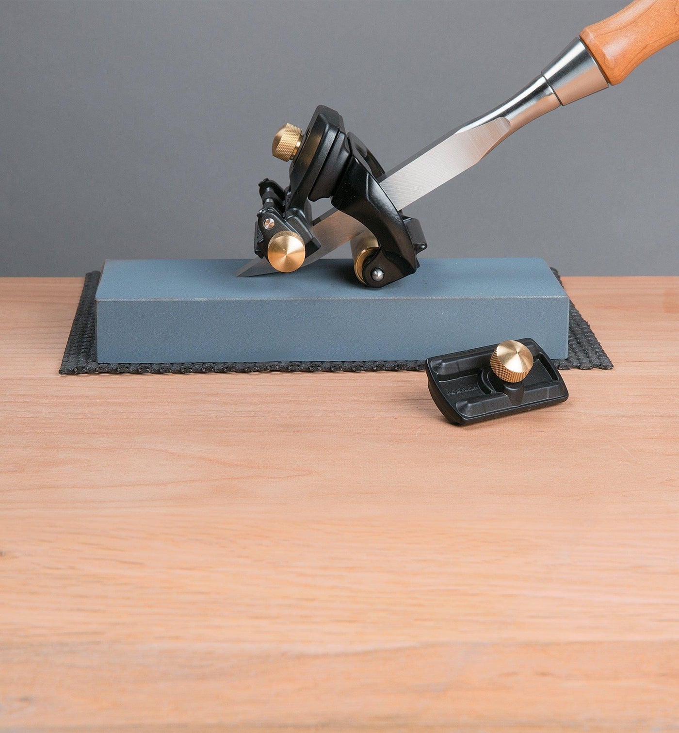 A VER Mortise Chisel Adapter for the Veritas Mk.II Narrow-Blade Honing Guide rests beside a chisel in a honing guide on a sharpening stone, all placed on a wooden surface.