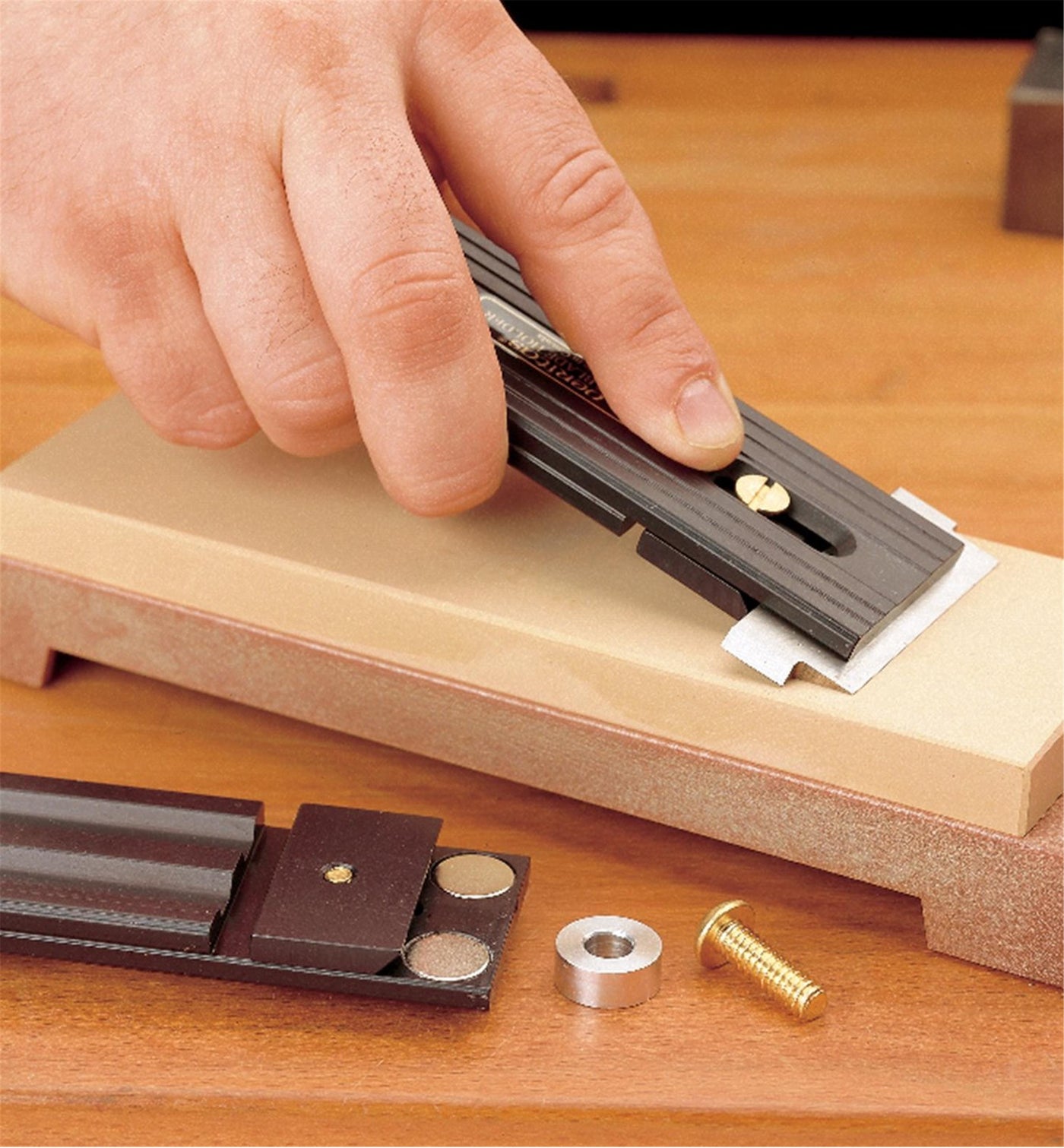 A person sharpens a chisel blade using the VER Veritas Small-Blade Holder, which features an anodized aluminum body, on a sharpening stone, with holder components and a brass screw resting on the wooden surface nearby.