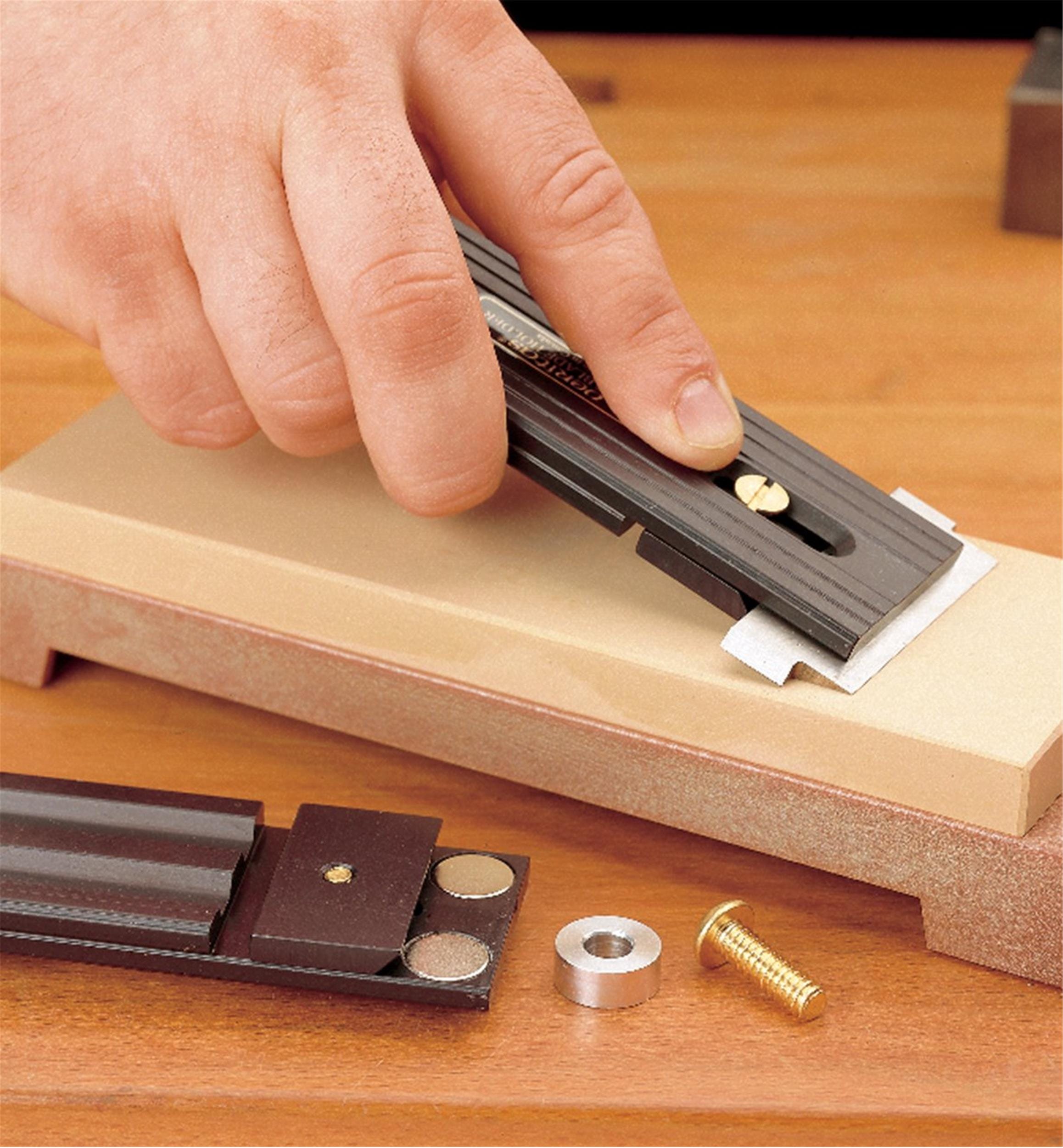 A person sharpens a chisel blade using the VER Veritas Small-Blade Holder, which features an anodized aluminum body, on a sharpening stone, with holder components and a brass screw resting on the wooden surface nearby.