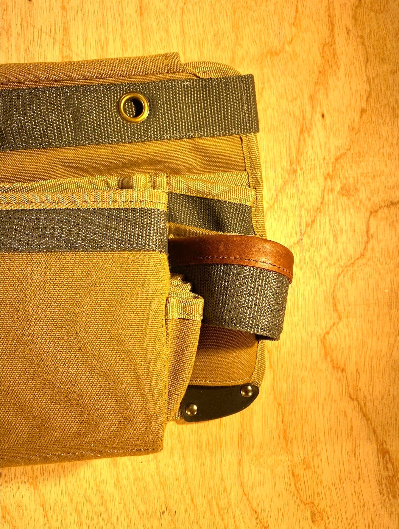 A close-up view of the Veto Pro Pac TA-WBX Waist Apron With Boxed Pockets by VET, made from durable tan and gray polyester canvas, featuring a leather accent and metal rivets, placed on a wooden surface.